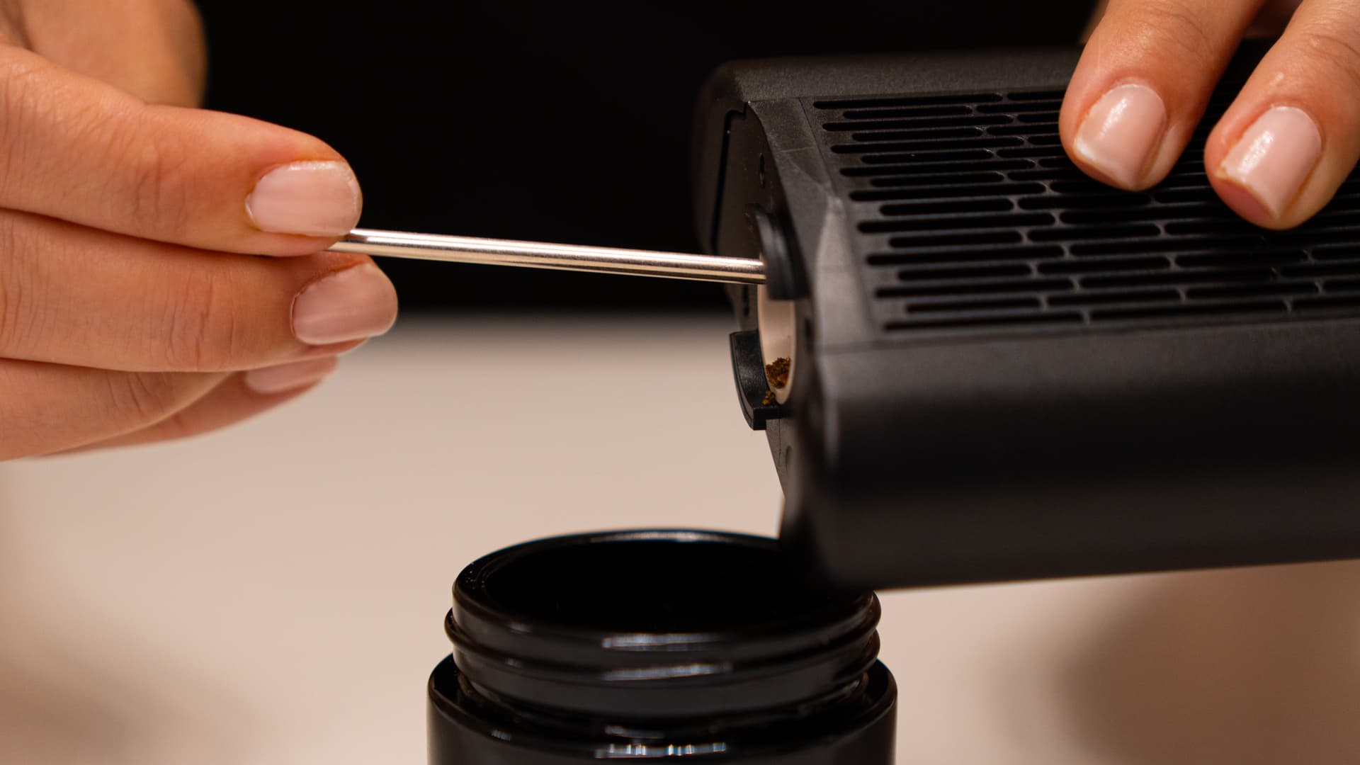 Hands holding the Angus Enhanced vaporizer inverted over a black collection container while using a stir tool to push spent herb out of the stainless steel oven chamber