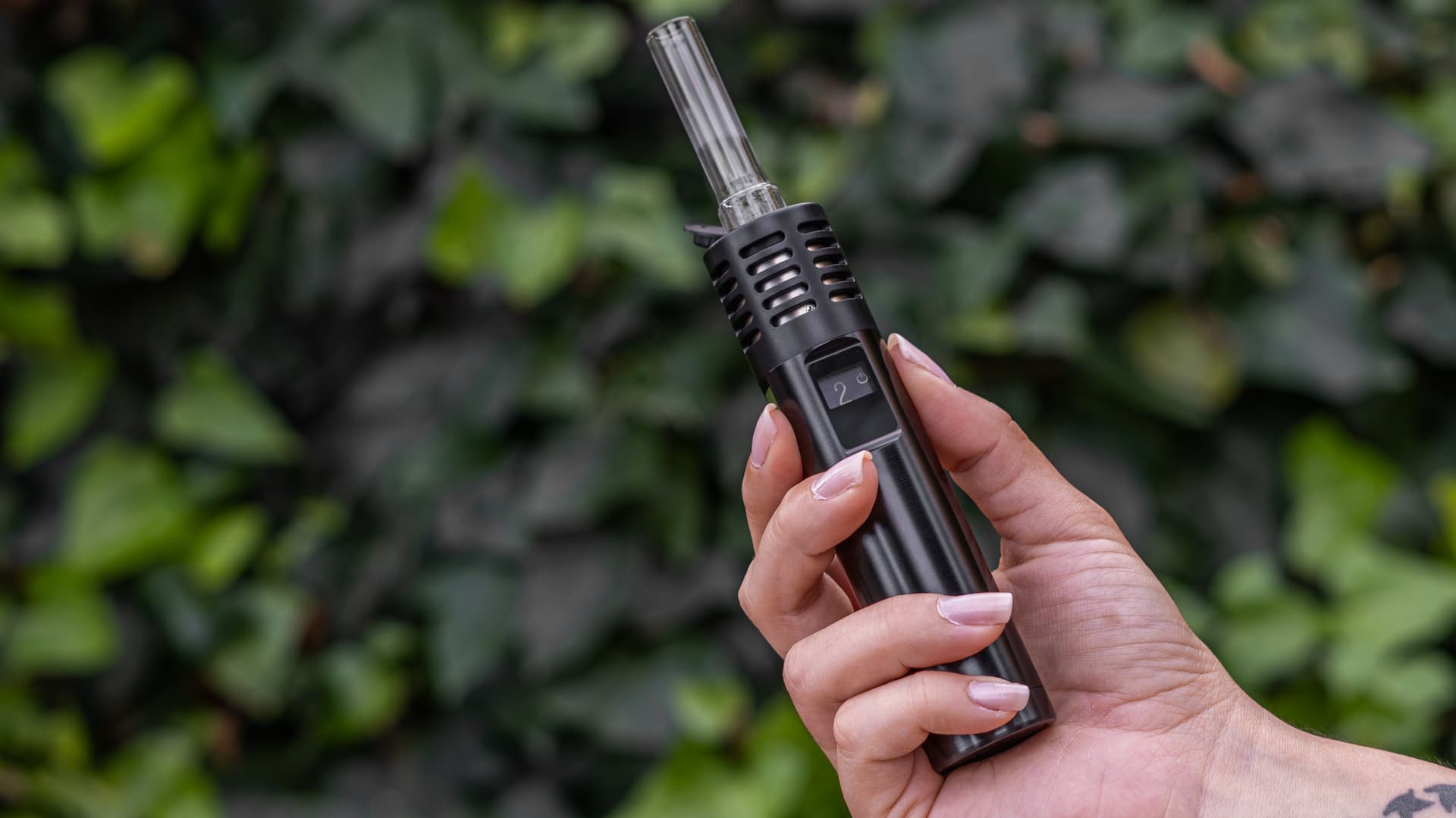 Arizer Air Max with glass aroma tube held in hand against green ivy backdrop showing portable form