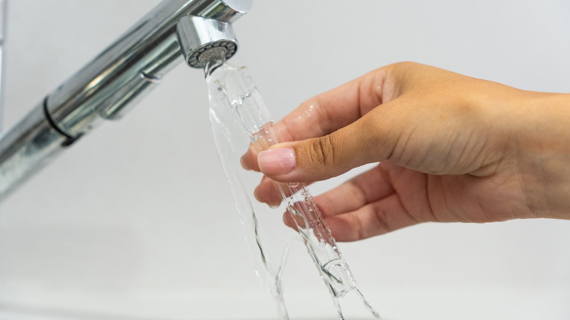 A glass aroma tube being rinsed under a chrome kitchen tap with water flowing through the clear tube