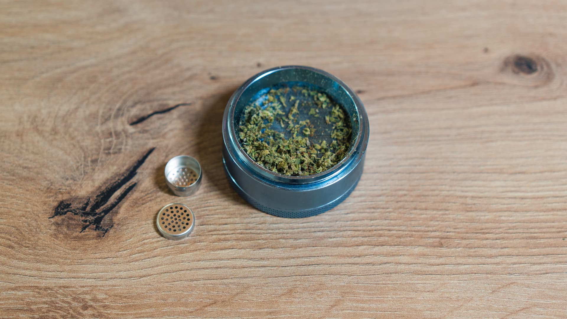 Storz & Bickel stainless steel dosing capsule and perforated screen lid next to a metal herb grinder filled with ground cannabis on a wooden surface
