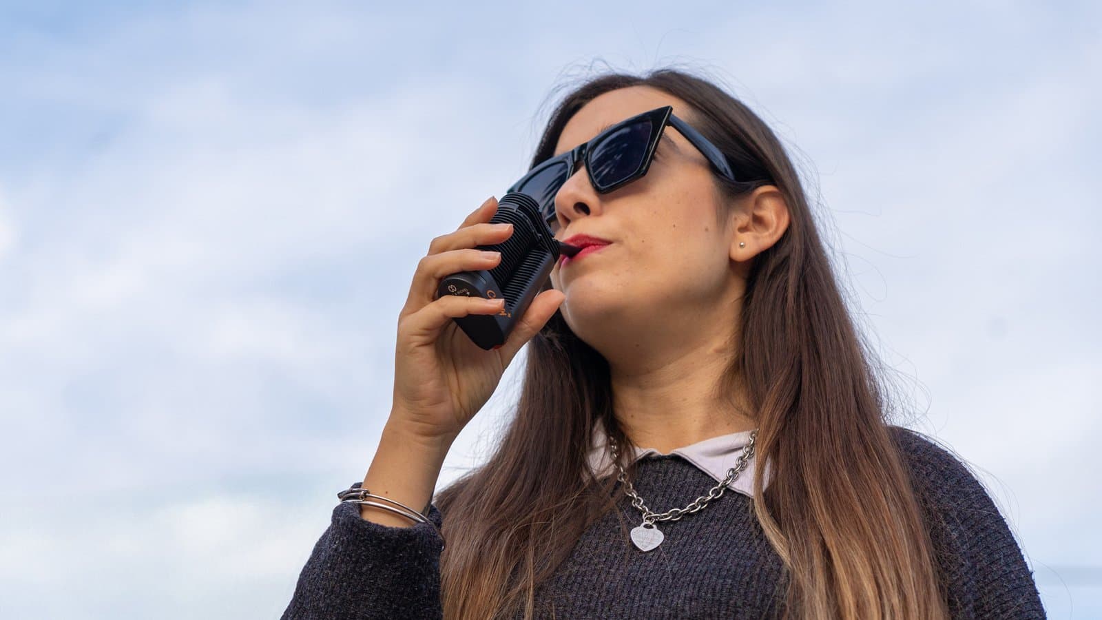 Frau mit Sonnenbrille dampft im Freien unter blauem Himmel aus einem schwarzen Crafty+