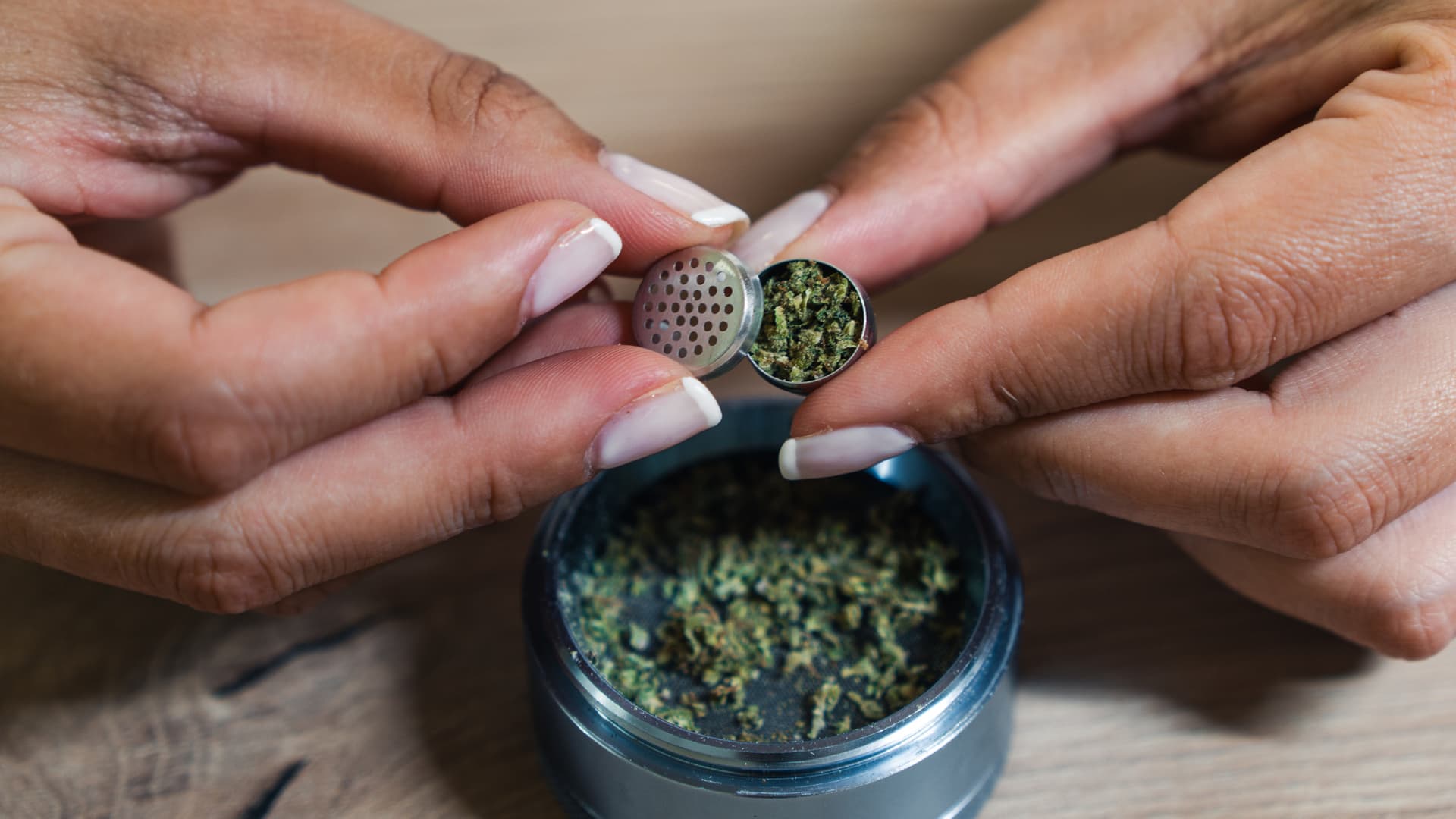A stainless steel dosing capsule loaded with ground cannabis held between fingers, with a 4-piece grinder full of material in the background.