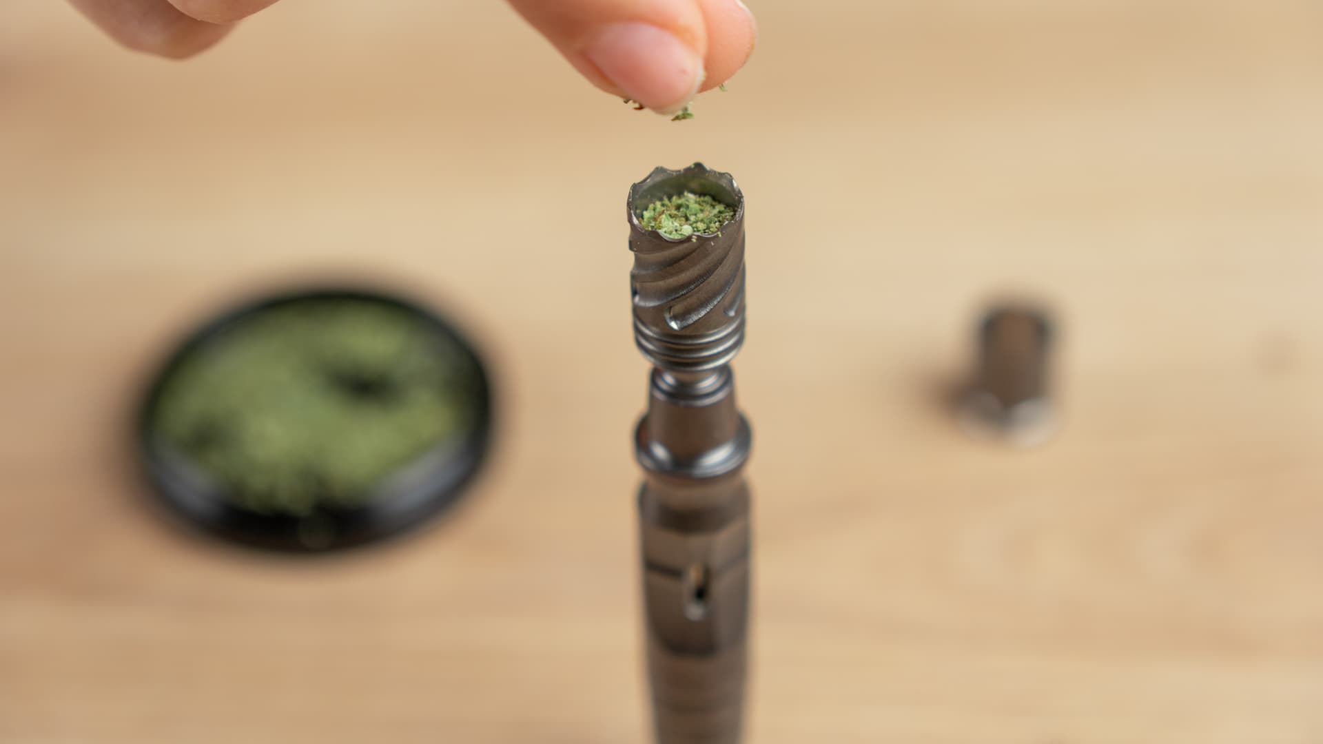 Hand füllt gemahlenes Cannabis in die Heizkammer des DynaVap HyperDyn Hyper Tip, Grinder im Hintergrund