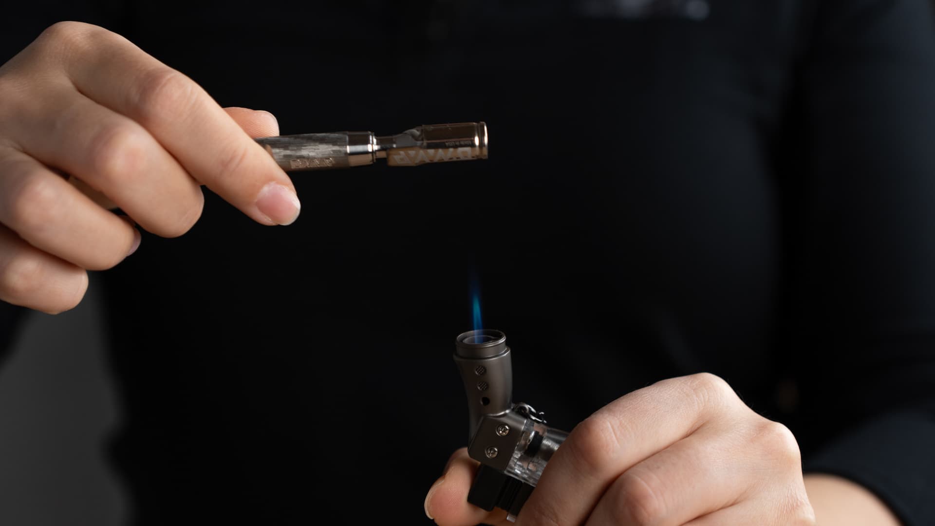 A DynaVap M7 held in hand and being heated by the blue flame of a butane torch, captured against a dark, moody background