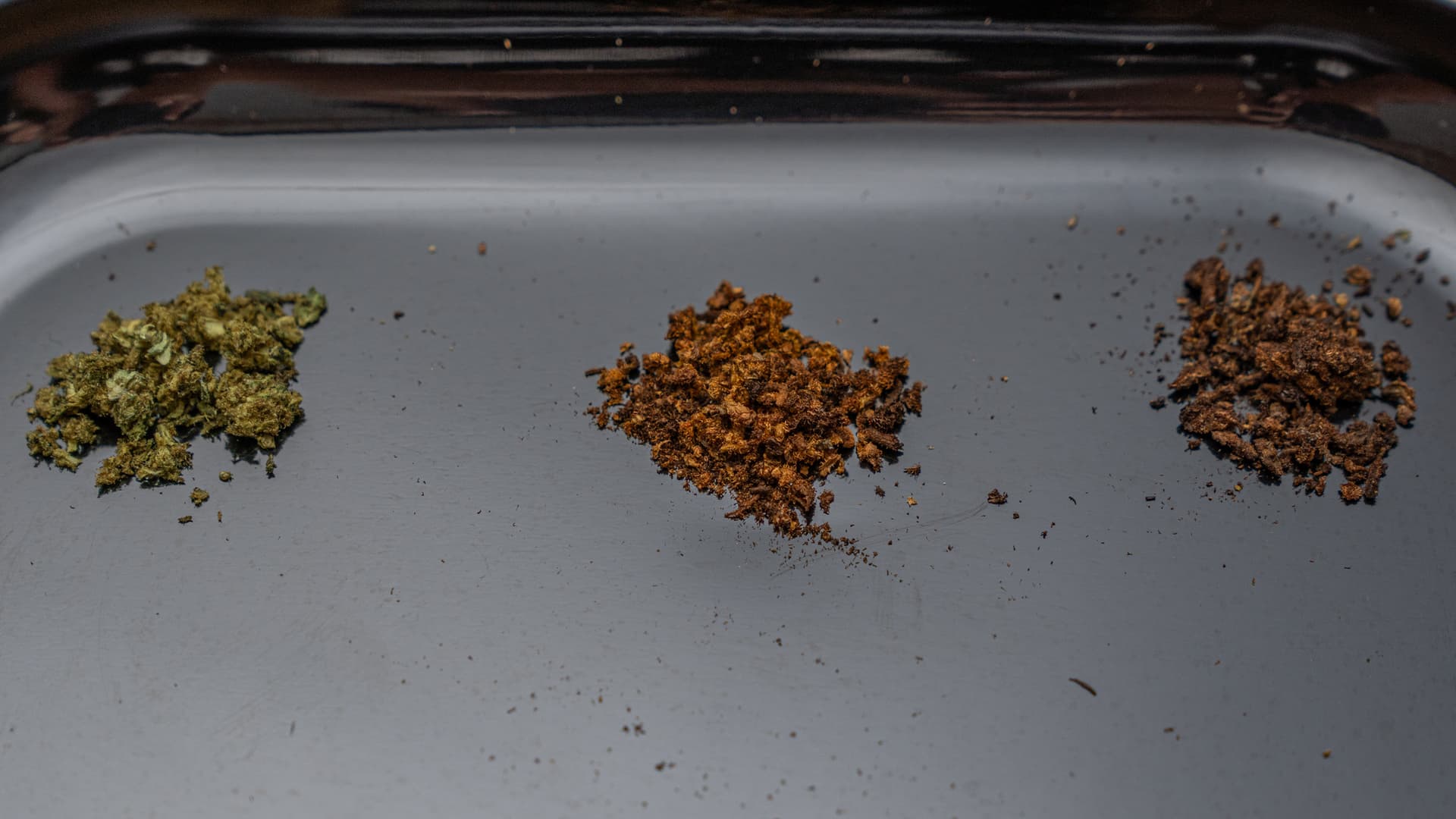 Three small piles of already-vaped cannabis on a dark slate tray, ranging from light golden-brown to medium amber-brown to dark chocolate, lit by soft natural light