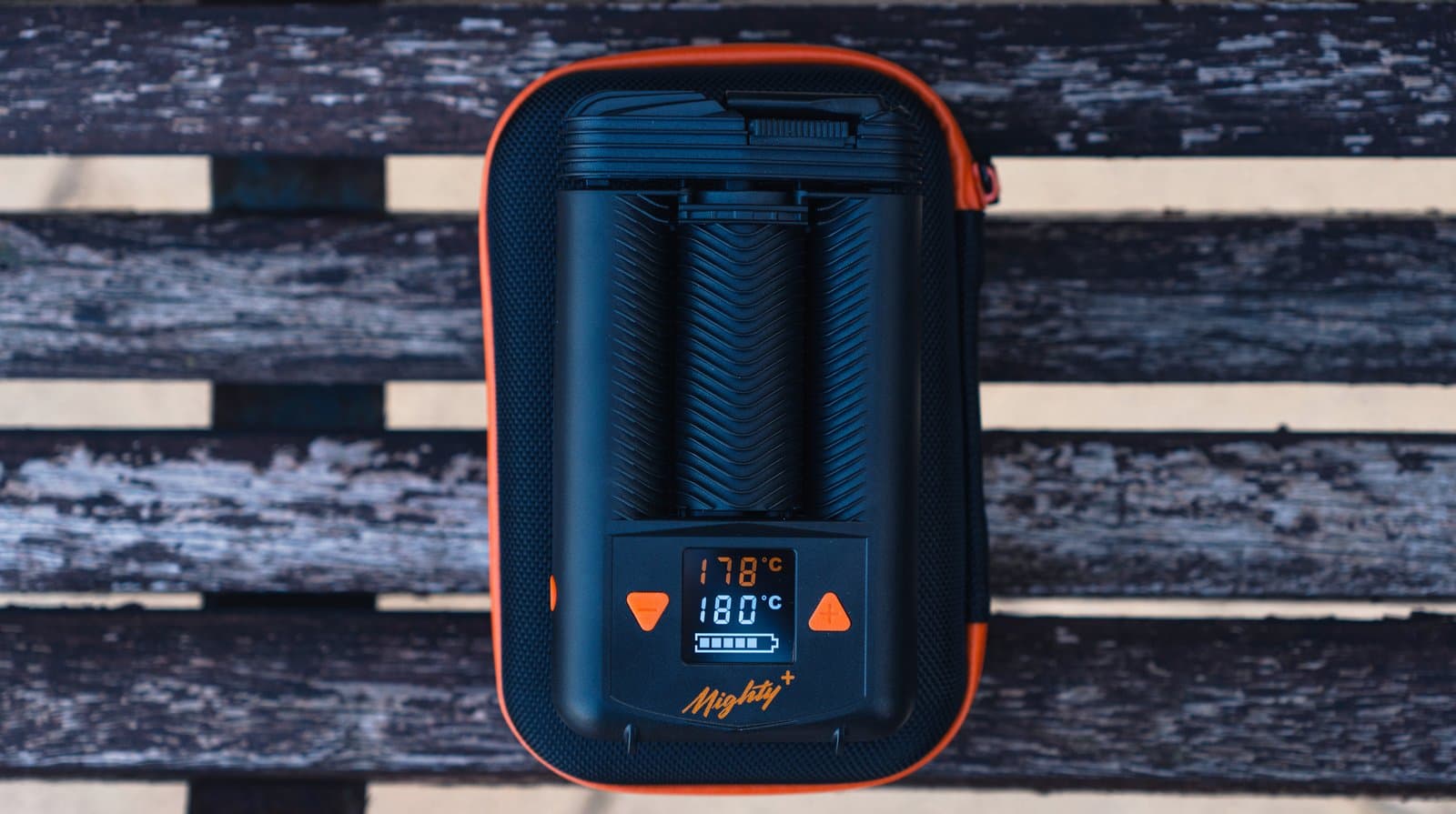 Schwarzer Mighty+ auf orangefarbenem Etui auf verwitterten Holzbrettern mit Heizanzeige im Display