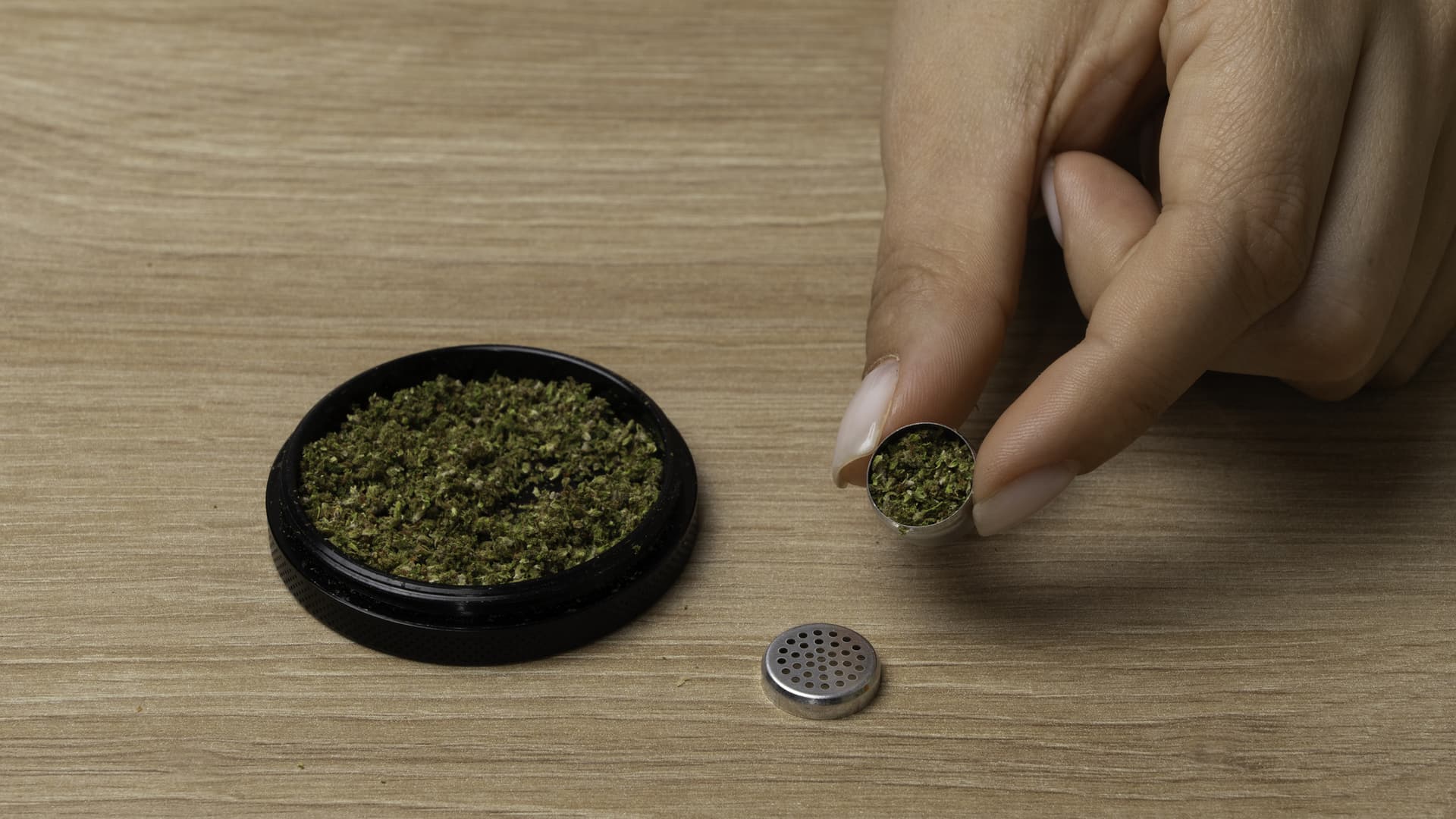 A dosing capsule loaded with ground cannabis sitting next to its screen lid on a wood surface