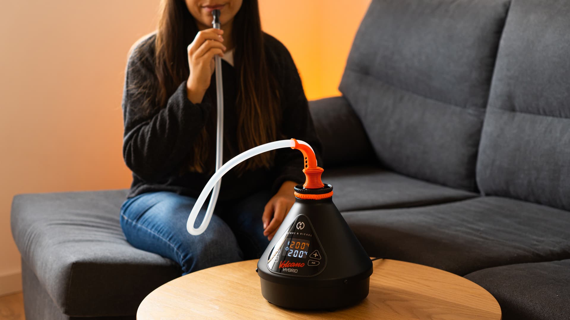 A person sitting on a sofa drawing from a Volcano Hybrid vaporizer placed on a wooden coffee table, in a softly lit living room with a warm peach-toned wall in the background