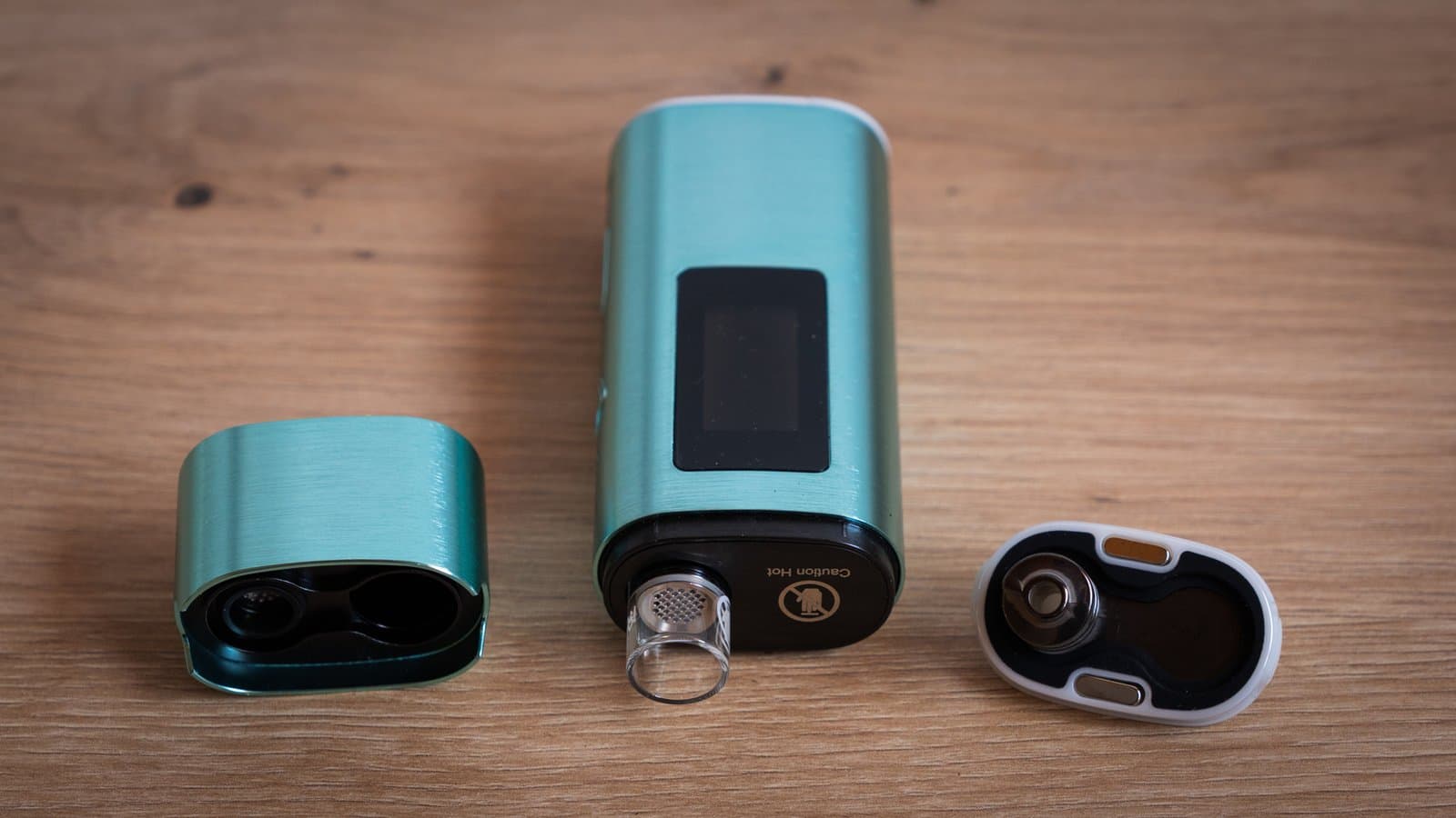 XLUX Roffu in Türkis zerlegt auf einer Holzoberfläche: magnetische Mundstückkappe mit freiliegendem Heizkammer-Sieb, Hauptgehäuse mit Glaskammer und Akkudeckel an der Unterseite