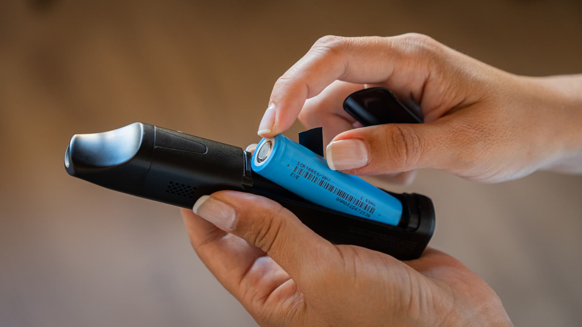 Hands sliding an 18650 lithium battery out of the bottom compartment of a portable vaporizer, with the knurled cap unscrewed and resting alongside