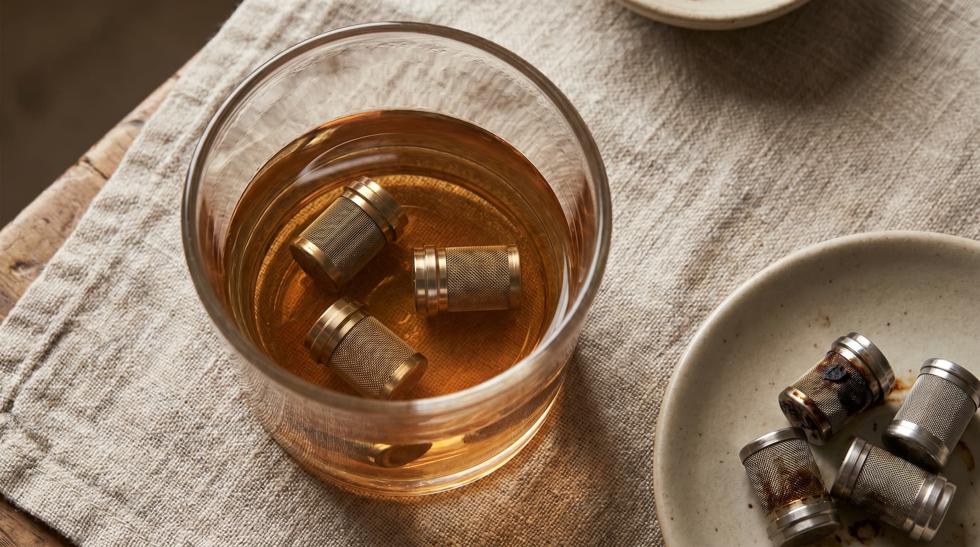 Drei Edelstahl-Dosierkapseln, vollständig in Isopropylalkohol eingetaucht, in einem durchsichtigen Trinkglas auf einem Leinentuch. Neben dem Glas liegen zwei benutzte Kapseln.