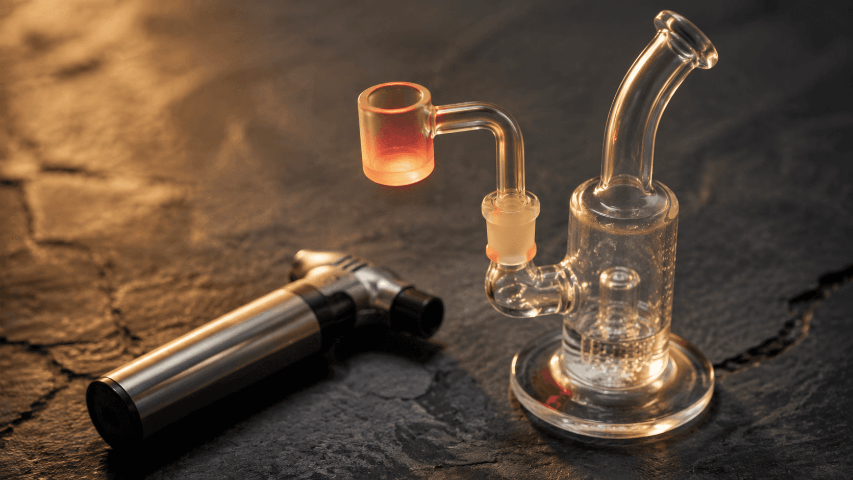 A heated quartz banger glowing faintly orange-red on a dark slate surface with a silver torch beside it