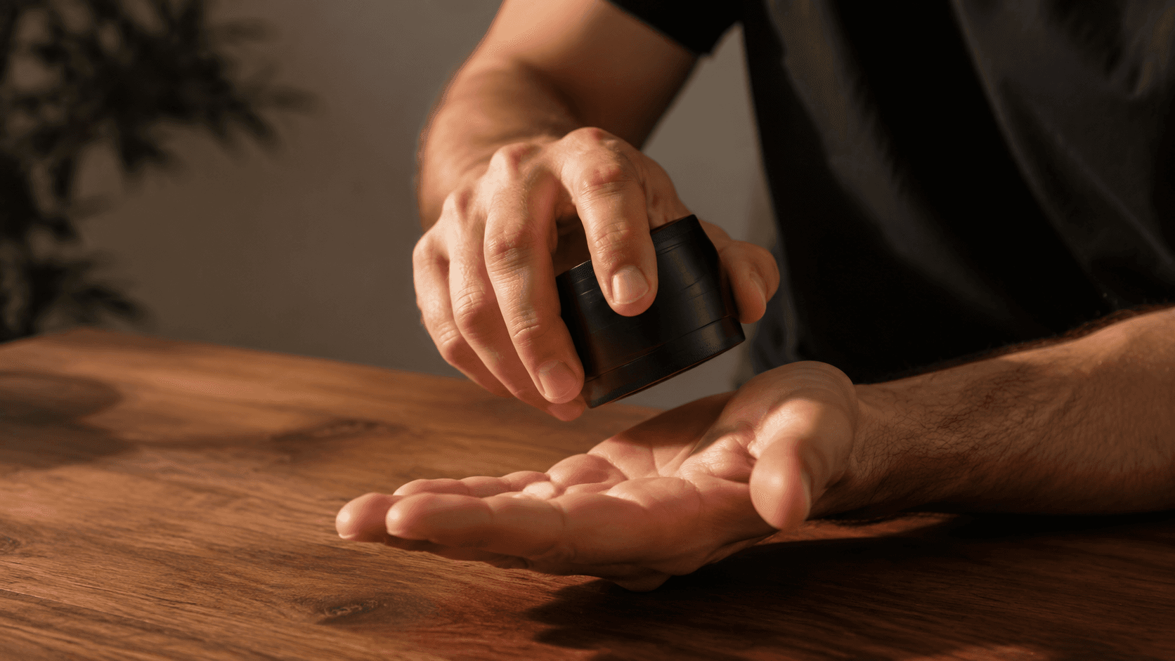 Hand tapping a closed matte black herb grinder against an open palm over a walnut wood table