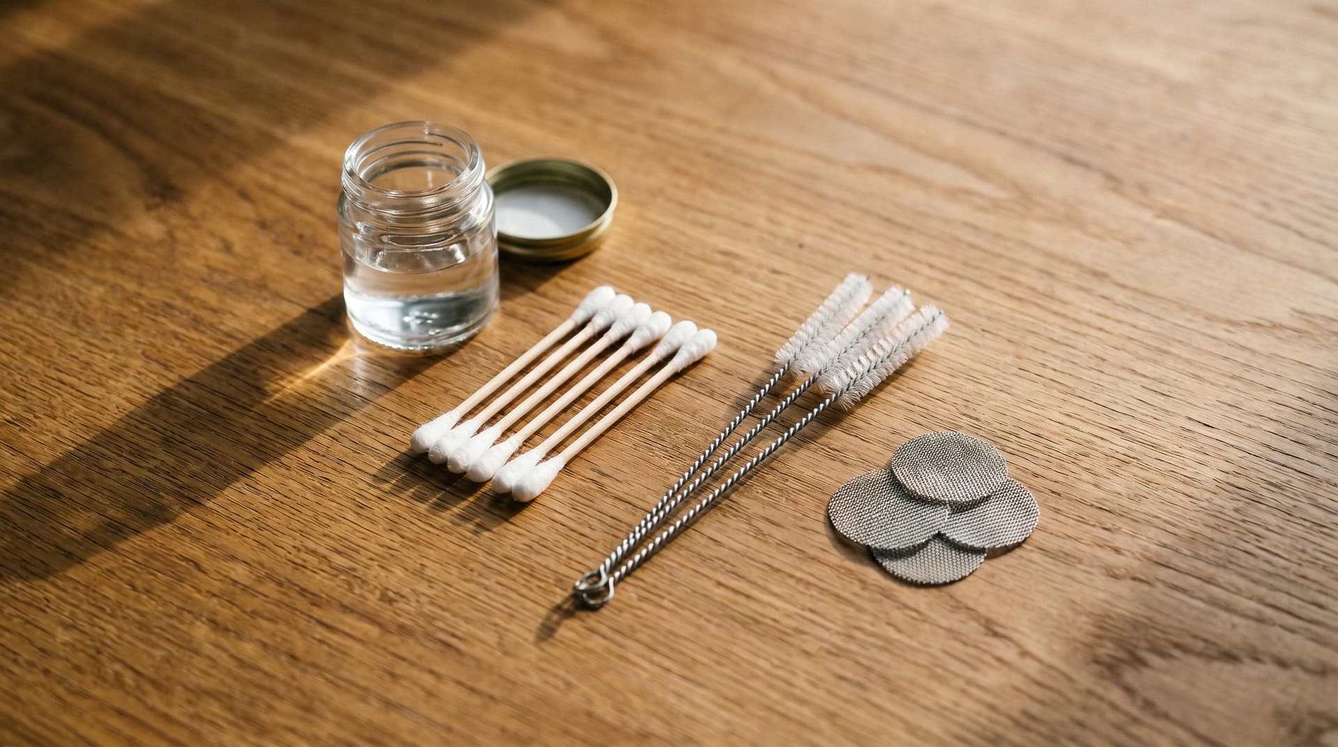 Overhead flat-lay on warm oak wood showing a small glass jar of isopropyl alcohol, a fanned row of cotton swabs, a bundle of three pipe cleaners, and a stack of round stainless steel replacement screens.