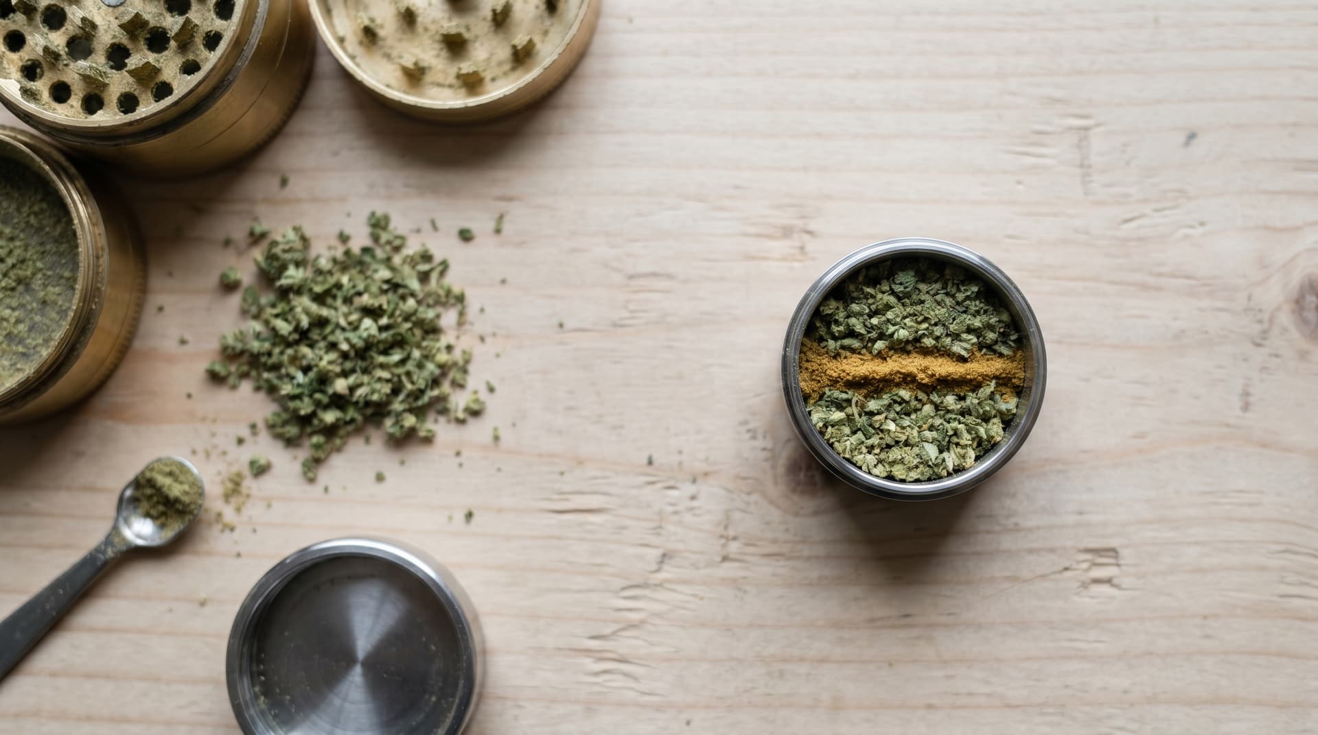 Macro overhead view of an open stainless steel dosing capsule on a light wood surface, showing three distinct layers inside: ground flower on the bottom, golden kief in the middle, and ground flower on top