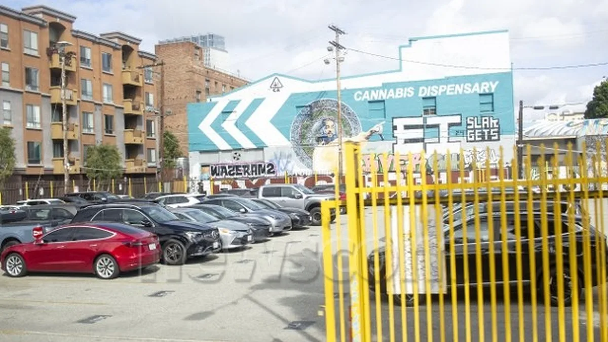 Cars fill a parking lot in front of a building with a mural reading "Cannabis Dispensary" behind a yellow fence in Los Angeles.