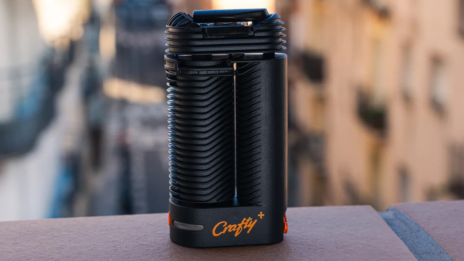 Black Crafty+ vaporizer with orange logo standing on a ledge against a blurred city street