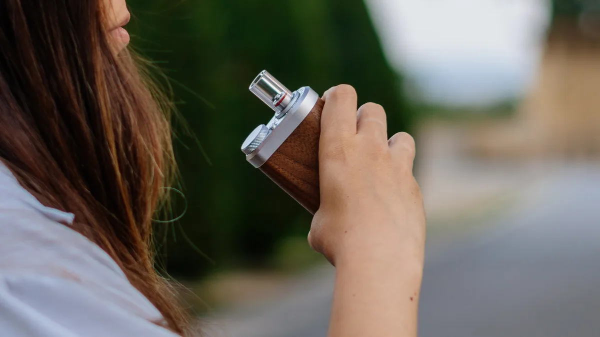 A person with long hair holds a wooden Tinymight 2 vaporizer with a metal mouthpiece near her face outdoors. | VapeExperts
