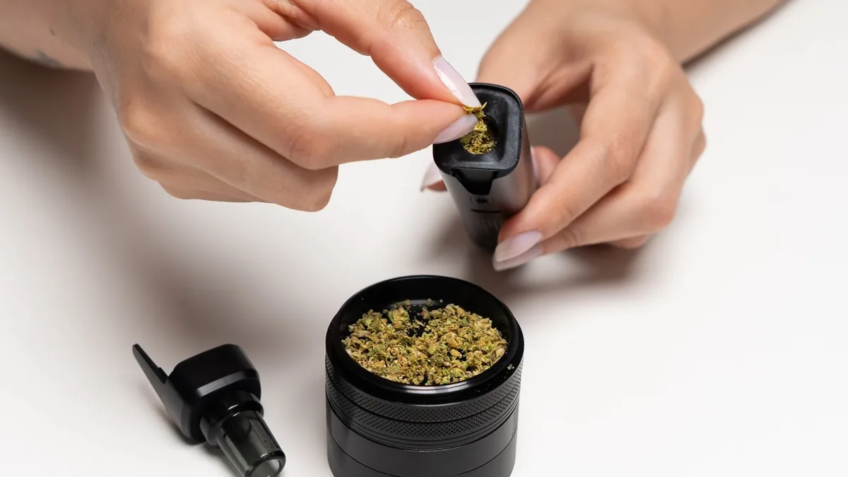 Hands loading ground cannabis into a black XMAX V4 Pro vaporizer above an open grinder on a white table. | VapeExperts