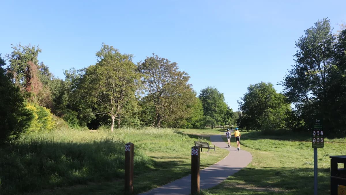 A winding paved path through green grass and trees in Cassiobury Park, with two people riding scooters in the distance under a clear blue sky.