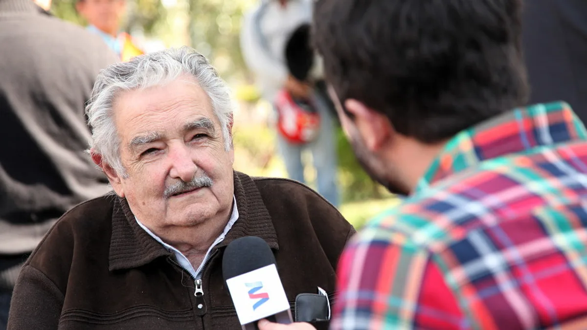 An elderly man with white hair and a mustache is interviewed outdoors by a reporter in a plaid shirt holding a microphone. | Bicentenario Uruguay/Wikimedia Commons (CC BY-SA 2.0)