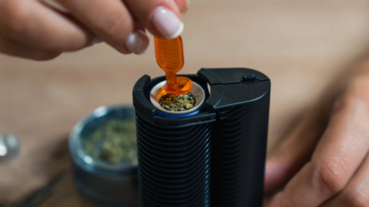 A hand uses a small orange tool to pack ground cannabis into the open chamber of a black portable vaporizer, with a grinder in the background. | VapeExperts