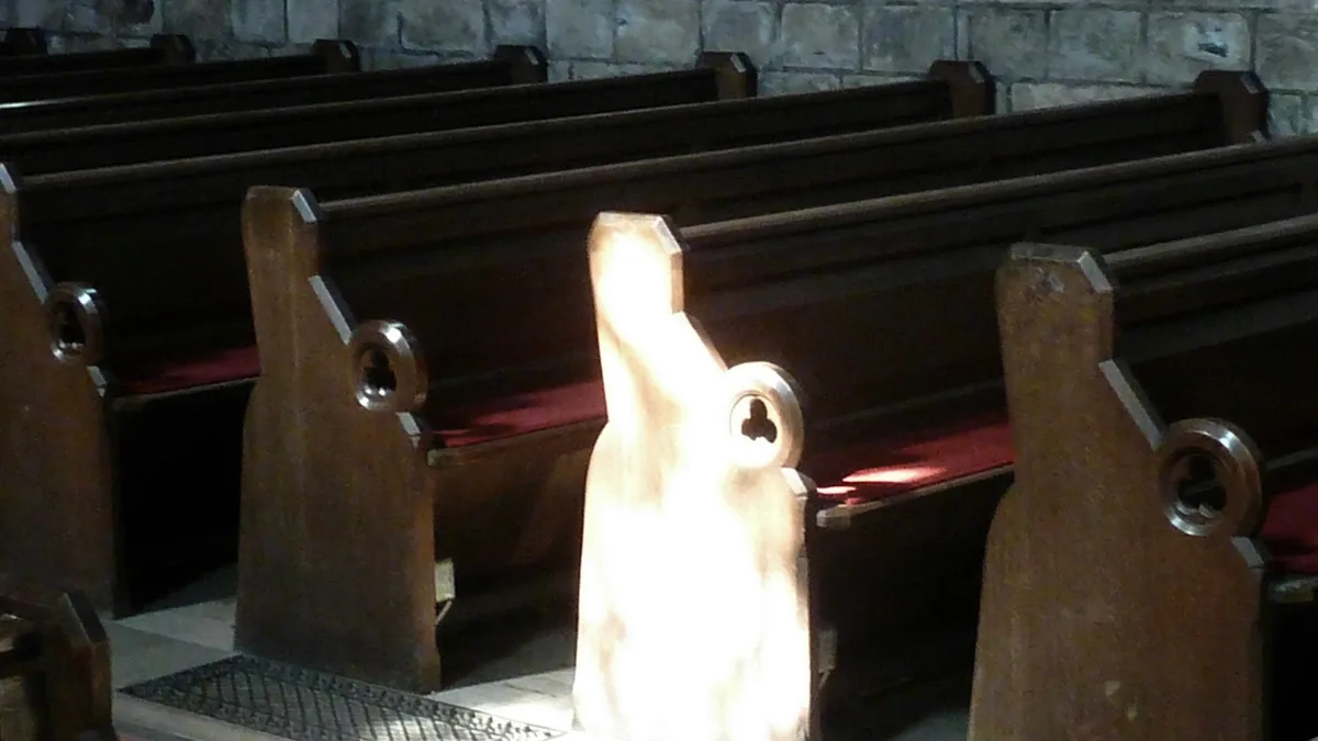 A beam of sunlight illuminates the end of a wooden church pew among rows of dark pews inside a stone church. | Storye book/Wikimedia Commons (CC BY-SA 4.0)