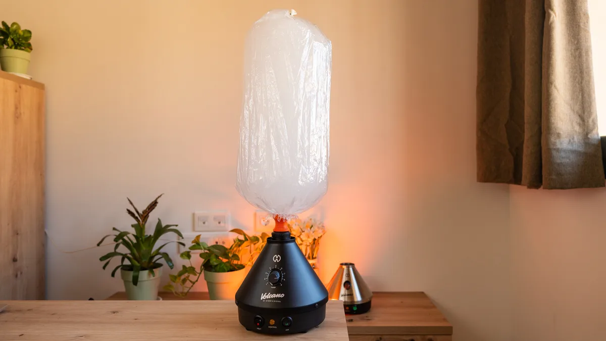 A black Volcano Classic vaporizer with a large inflated clear balloon bag attached, standing on a wooden table near potted plants. | VapeExperts