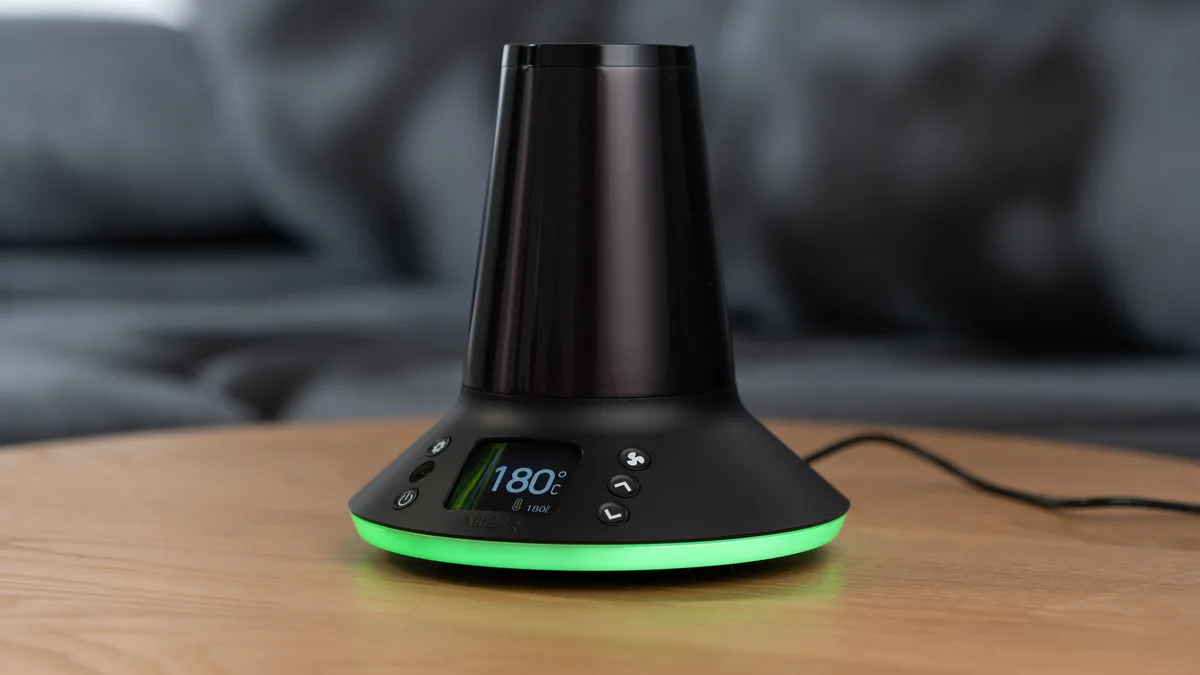 A black Arizer XQ2 desktop vaporizer with a glowing green base sits on a wooden table in front of a gray couch. | VapeExperts