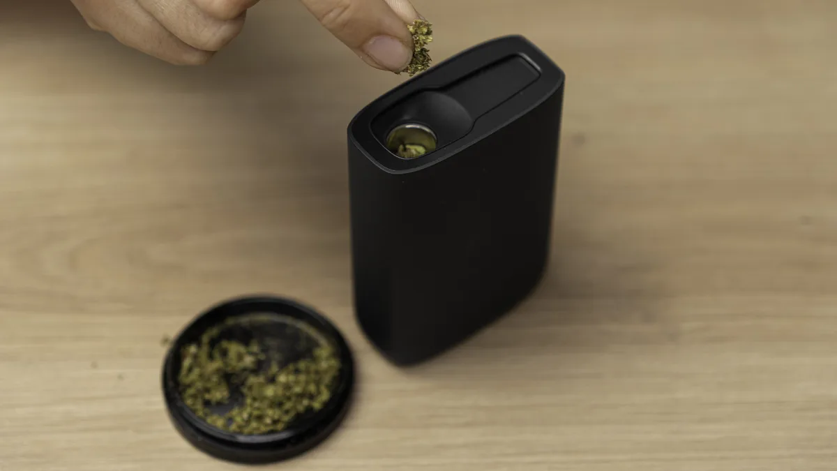 A hand drops ground cannabis into the open chamber of a black Fenix NEO vaporizer, with a grinder lid holding herb on a wooden table. | VapeExperts