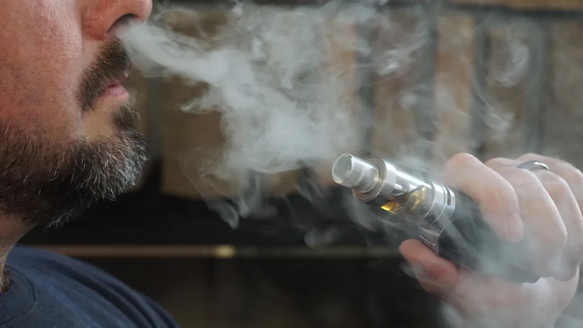 A bearded man exhales a thick cloud of vapor while holding a silver e-cigarette tank device in his hand. | Vaper City/Wikimedia Commons (CC BY-SA 4.0)