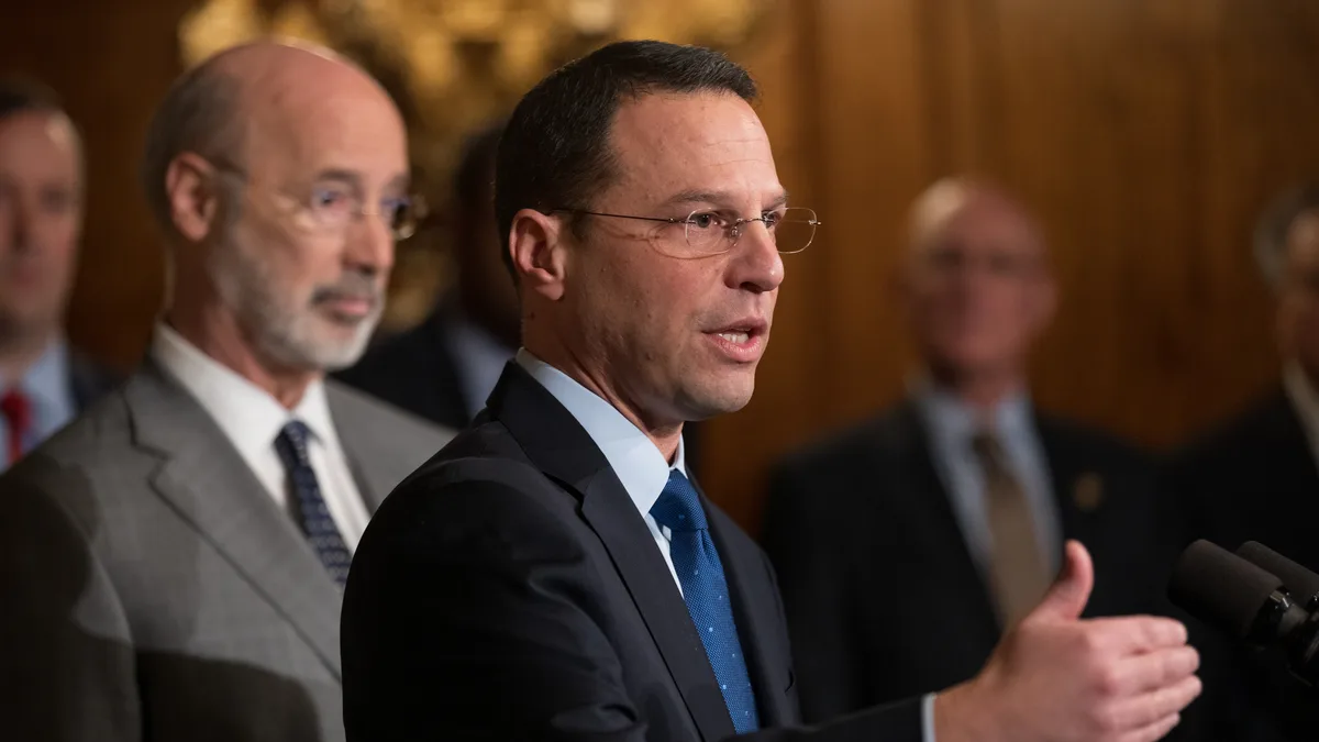 A man in glasses and a dark suit speaks at microphones while an older man in a gray suit stands behind him. | Governor Tom Wolf from Harrisburg, PA/Wikimedia Commons (CC BY 2.0)