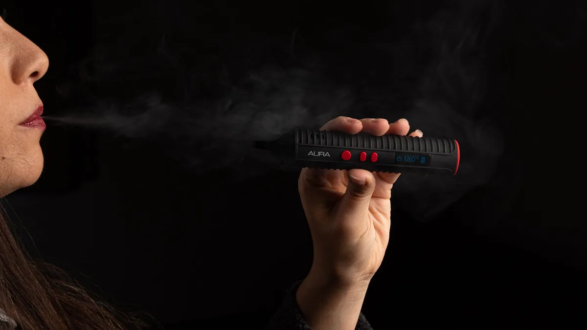 A woman exhales a cloud of vapor while holding a black Flowermate Aura vaporizer against a dark background. | VapeExperts
