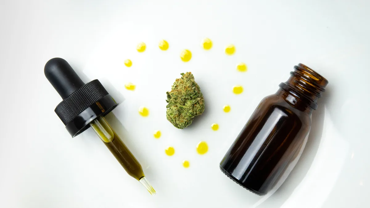 A cannabis bud on a white plate surrounded by yellow oil droplets, with a filled dropper on the left and an open amber glass bottle on the right. | Sherpa SEO/Wikimedia Commons (CC BY 3.0)