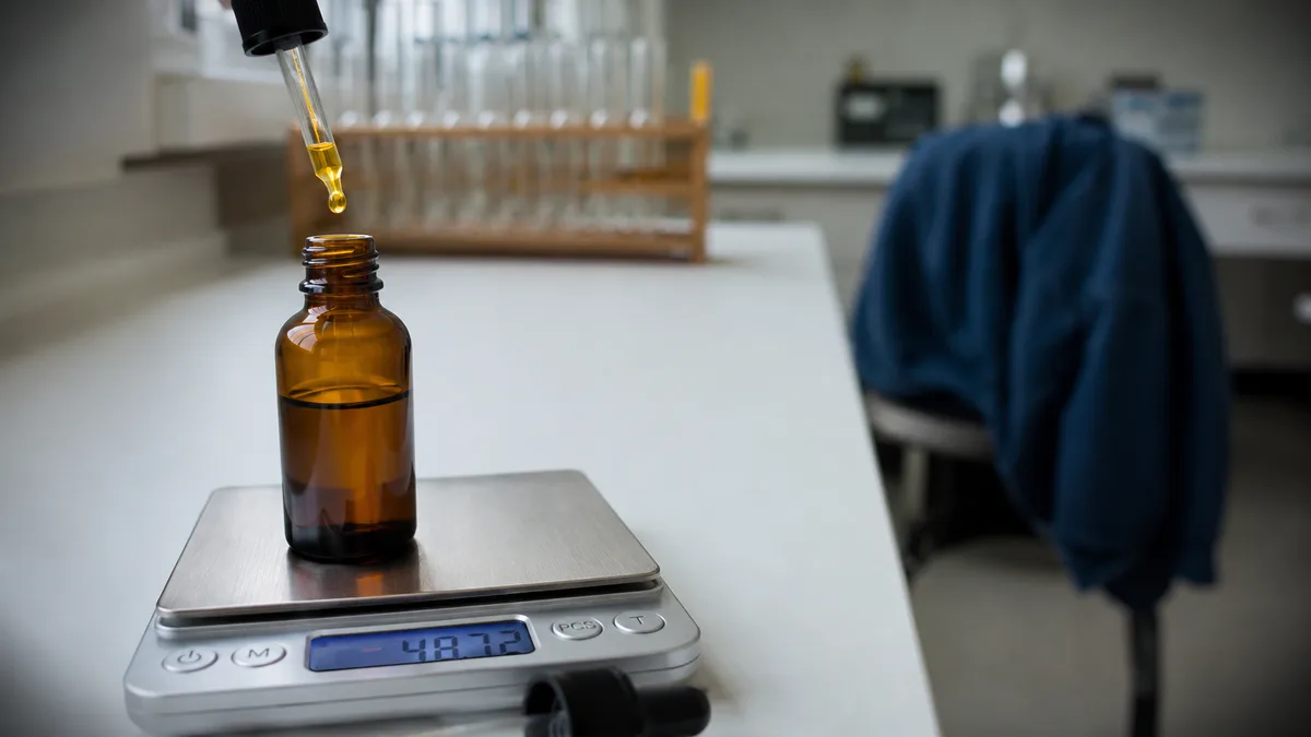 Amber glass dropper bottle of CBD oil beside a small digital kitchen scale on a clean white countertop, no visible labels or text
