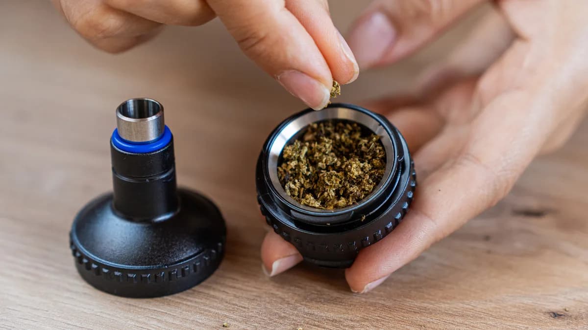 Fingers sprinkle ground herb into the metal filling chamber of a black vaporizer part held over a wooden table. | VapeExperts
