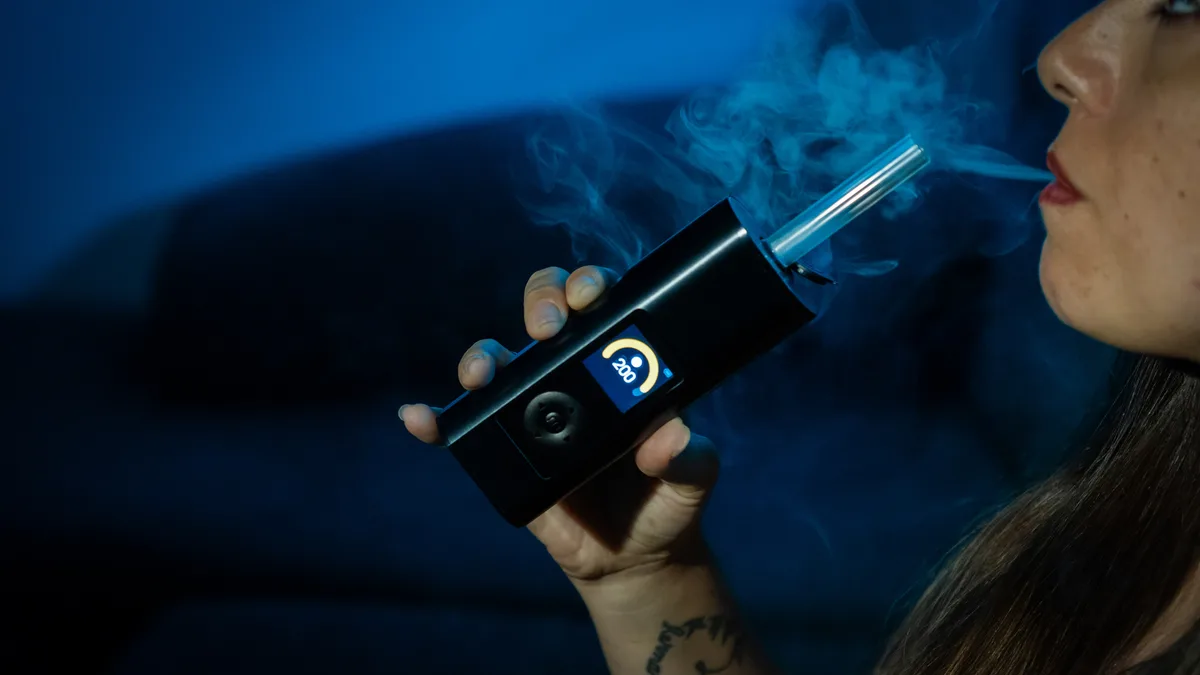 A woman exhales vapor while holding a black Arizer Solo 3 vaporizer with a glass stem to her lips in a dimly lit room. | VapeExperts