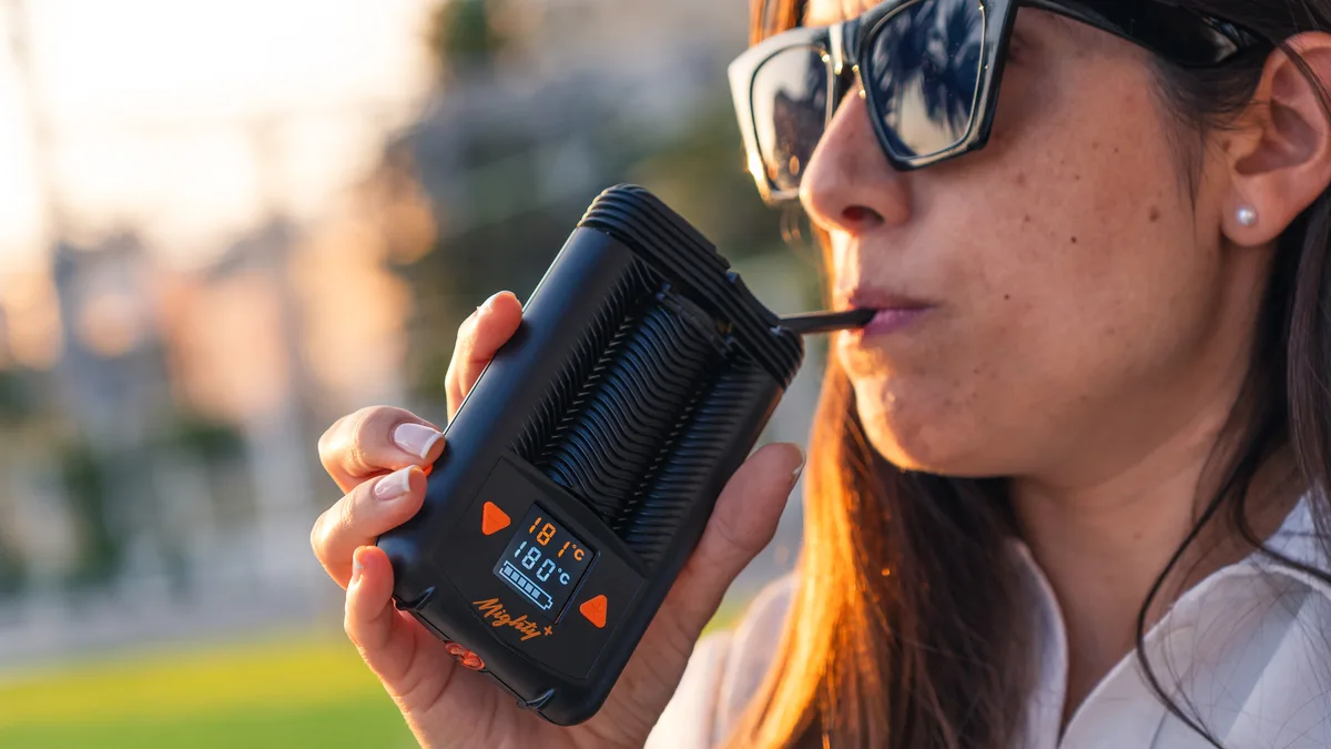 A woman wearing sunglasses inhales from a black handheld Mighty+ vaporizer held to her mouth outdoors. | VapeExperts