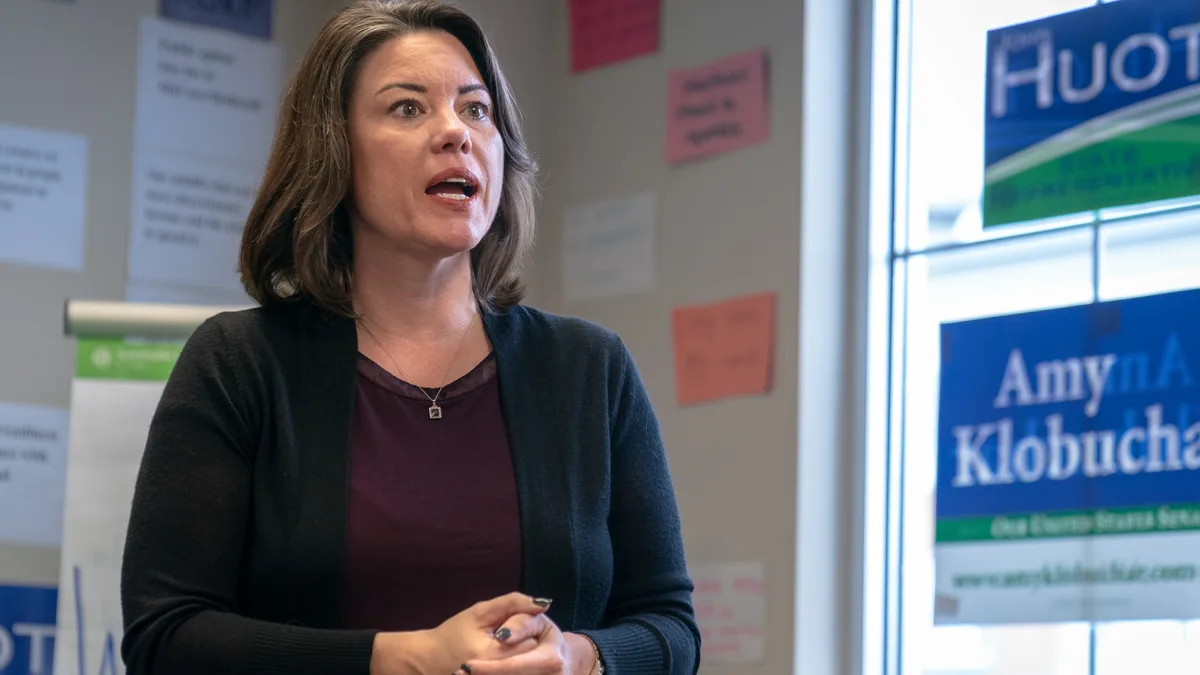 Angie Craig speaking indoors in front of a window with campaign signs, wearing a dark cardigan. | Lorie Shaull/Wikimedia Commons (CC BY-SA 2.0)