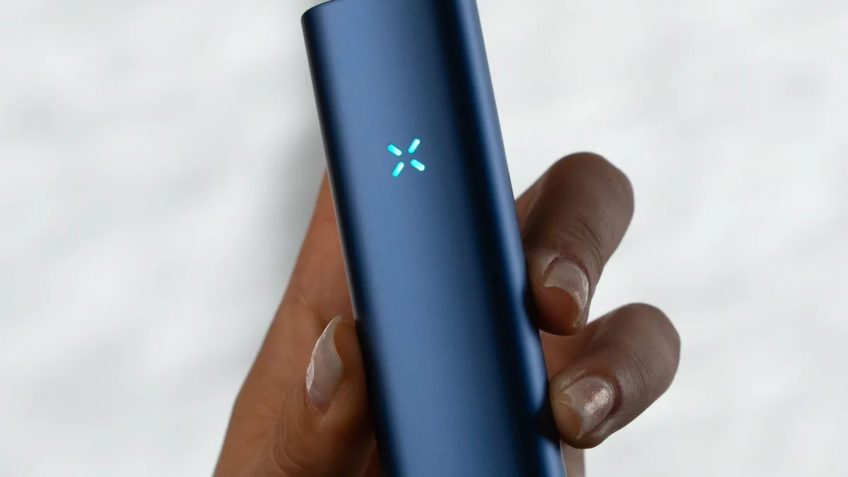 A hand holds a dark blue PAX Plus vaporizer with a glowing X-shaped light on its face against a blurred white background. | VapeExperts