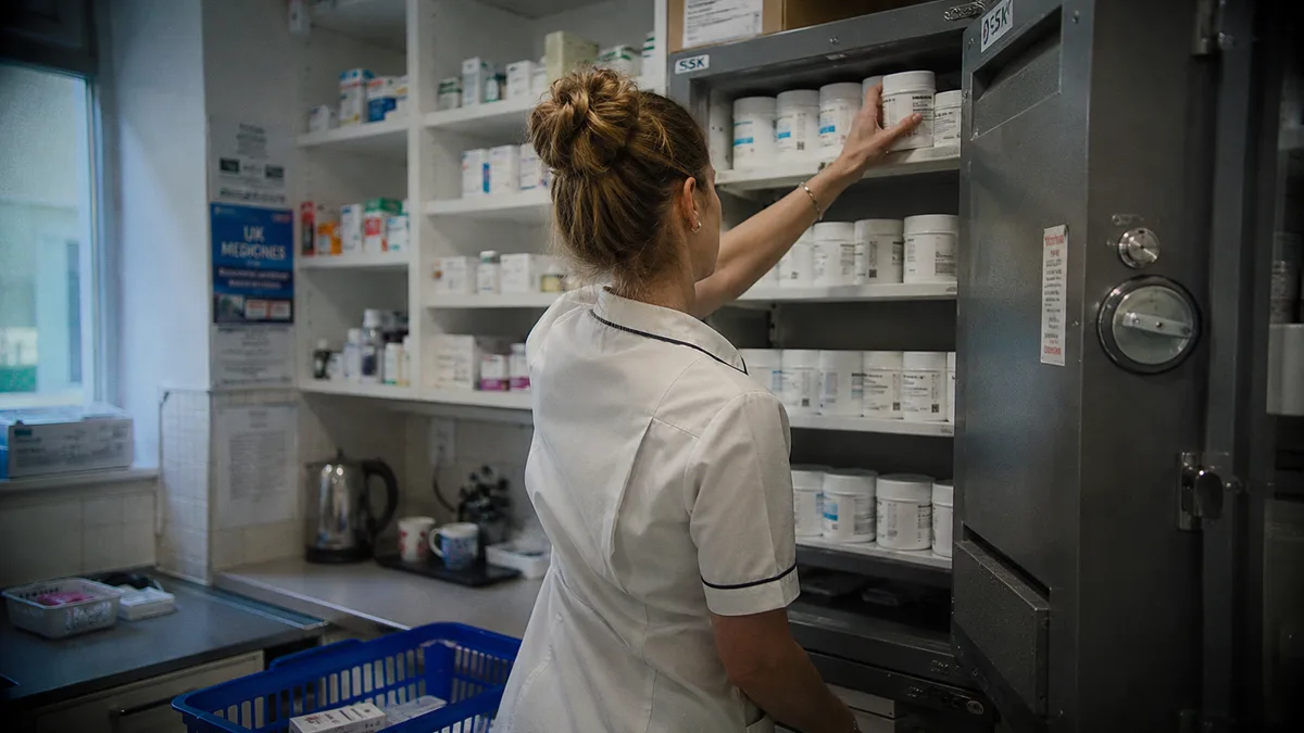 Pharmacist in a private pharmacy reaching into a locked controlled-drugs cabinet, unmarked white medication tubs on the shelf, no legible labels visible.