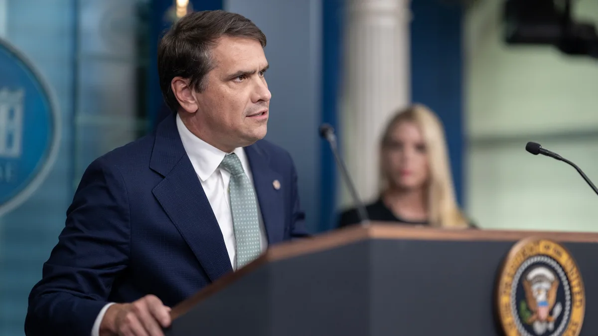 A man in a dark suit speaks at a podium bearing the presidential seal, with a blurred woman in the background. | The White House/Wikimedia Commons (Public domain)