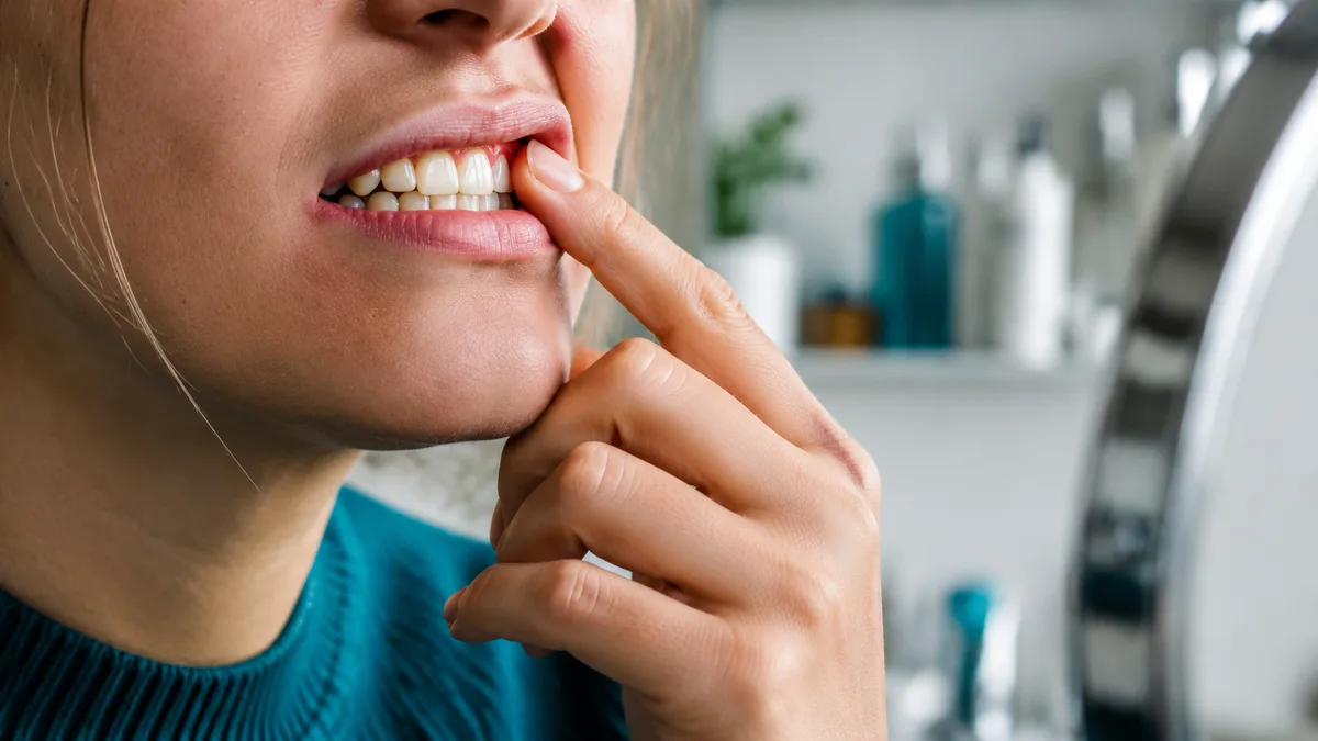 Dentist in gloves using a mouth mirror to examine a patient's gum line under an exam light, tight crop on hands and mouth.