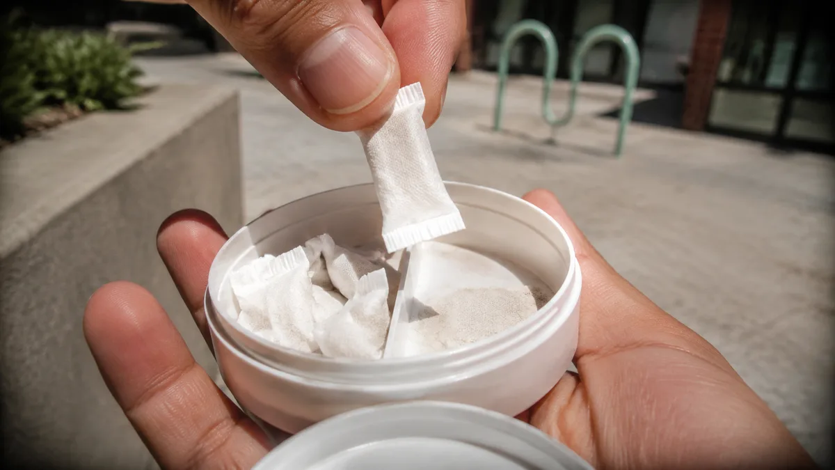 Close-up of fingers lifting a small white pouch from an open, unmarked round child-resistant puck with fresh and used compartments visible.