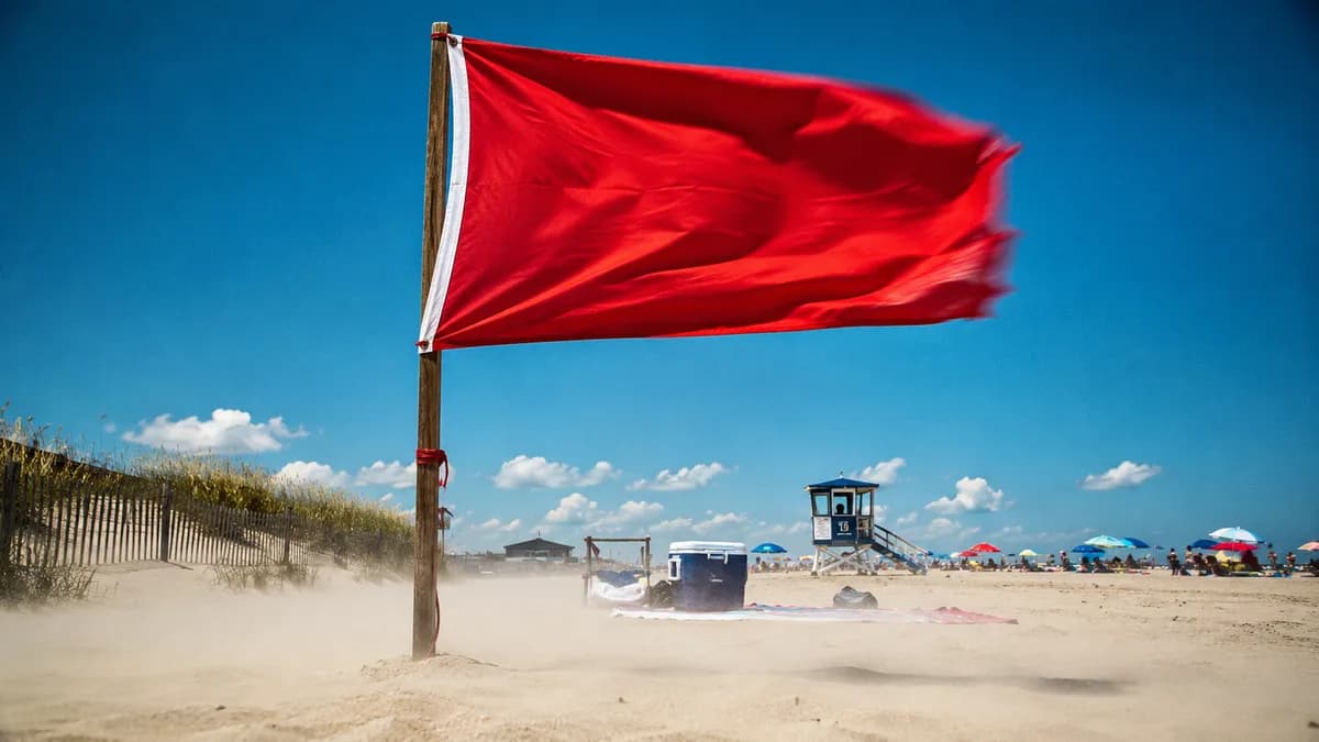 A red beach warning flag snapping taut in strong onshore wind, sand streaming low across the ground behind it, overcast bright sky.