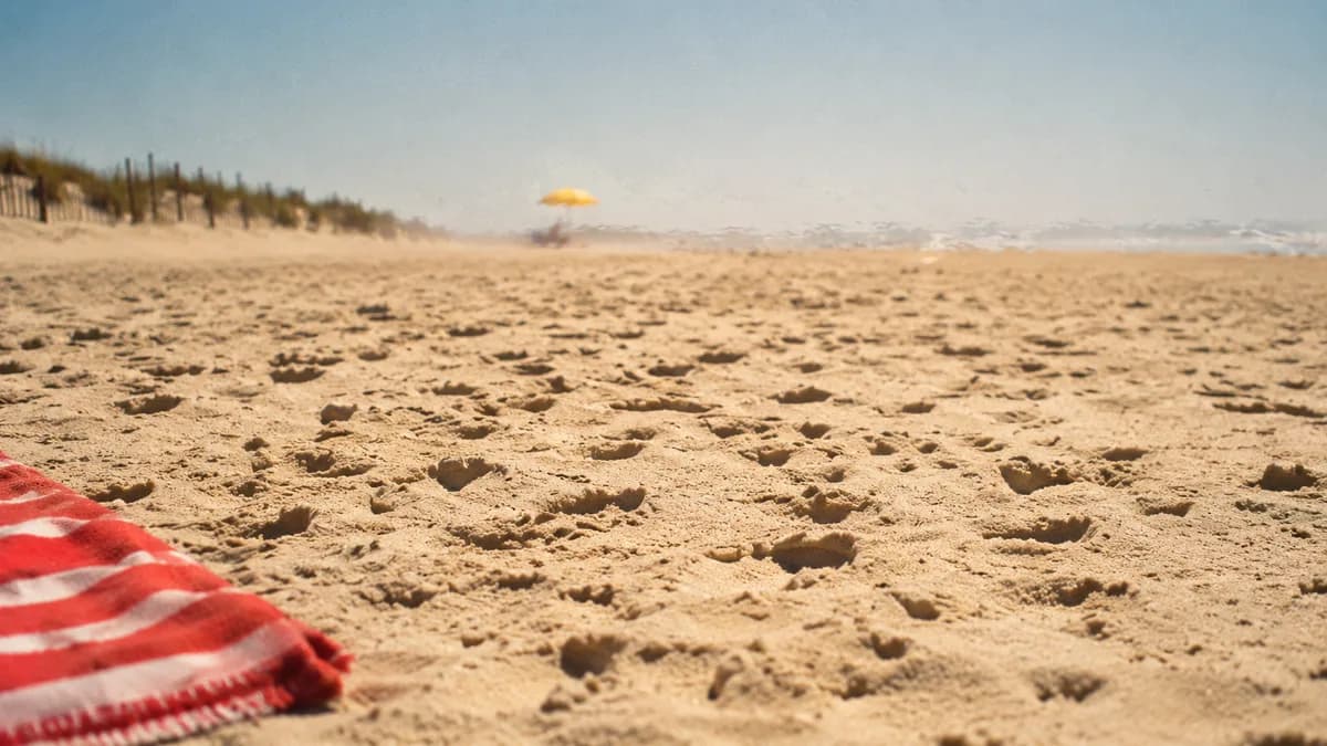 Sun-baked beach sand in harsh midday light, bare footprints and the edge of a towel, visible heat shimmer rising off the surface.