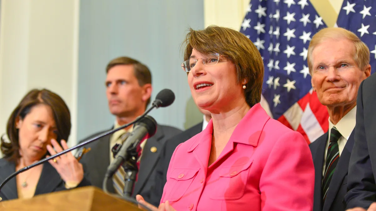 A woman in a pink jacket speaks into microphones at a podium while other officials stand behind her in front of American flags. | Senate Democrats/Wikimedia Commons (CC BY 2.0)