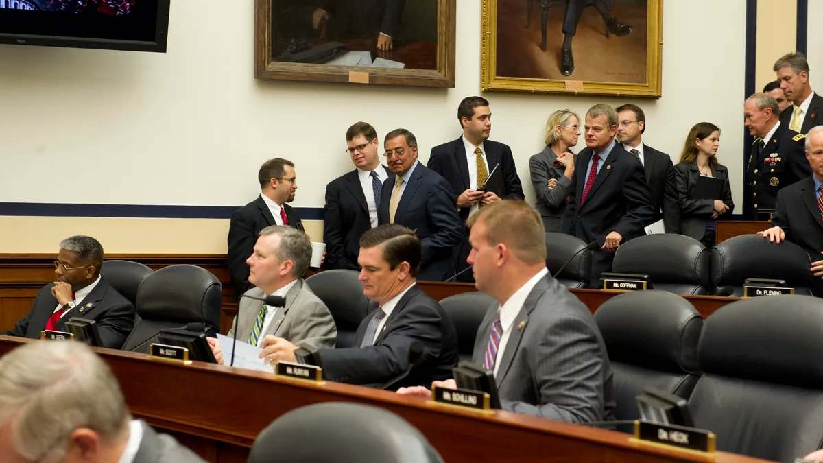Men in suits sit at tiered wooden desks with nameplates in a congressional hearing room while others stand talking behind them. | Erin Kirk-Cuomo/Wikimedia Commons (Public domain)