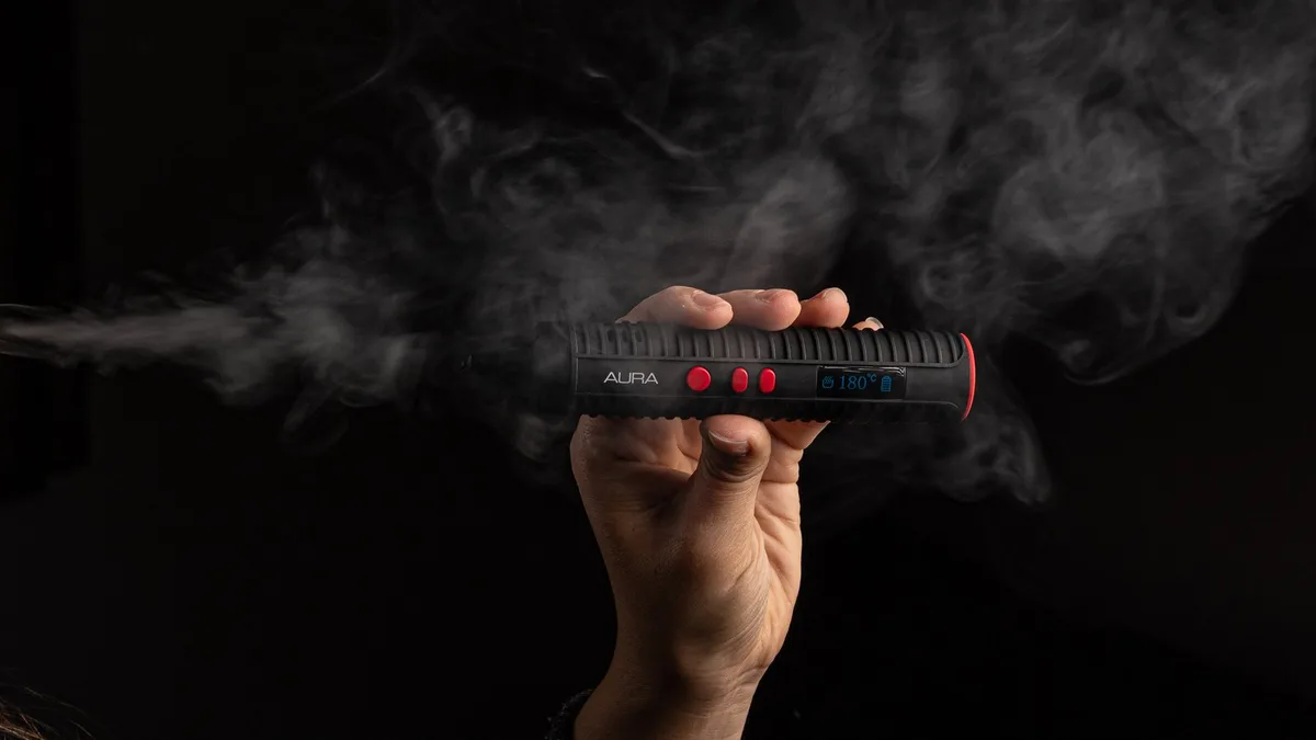 A hand holds a black Flowermate Aura vaporizer surrounded by vapor against a dark background. | VapeExperts