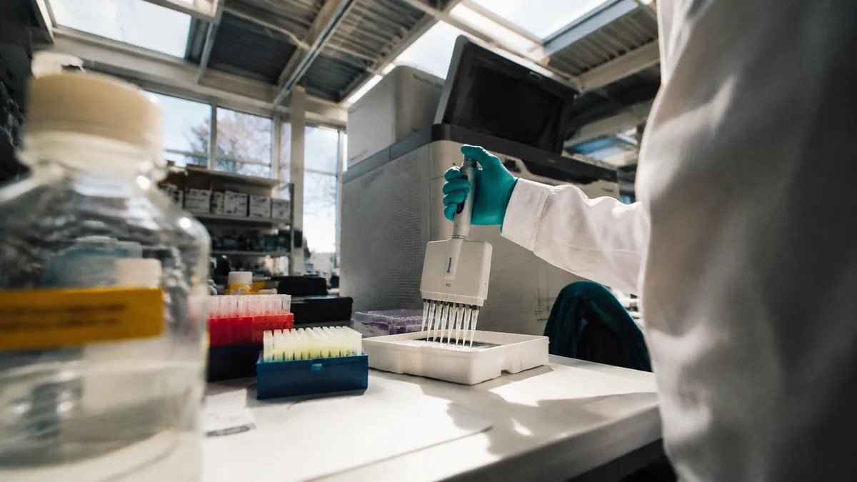 Gloved technician pipetting samples into a tray beside a DNA sequencing machine in a genomics lab, no legible screens or labels in focus.